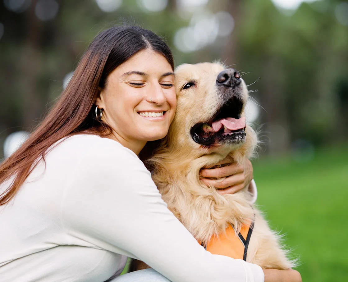 woman sits next to and hugs a golden retriever outside in park