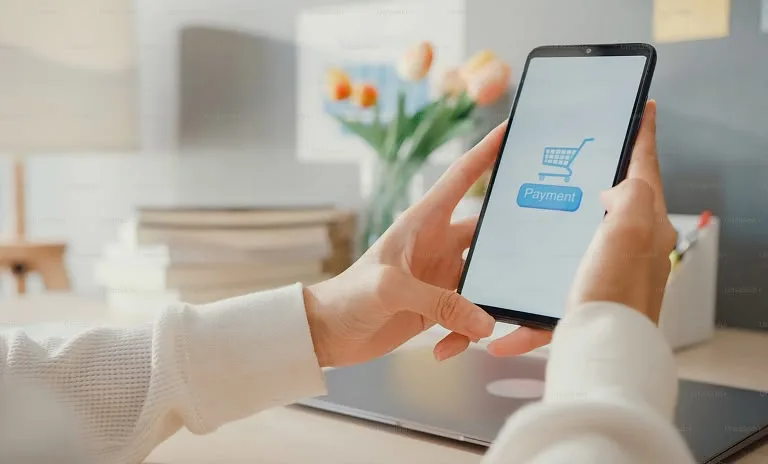 A person holds a smartphone displaying a shopping cart icon and a blue Payment button, at a bright wooden desk with a laptop and tulips.