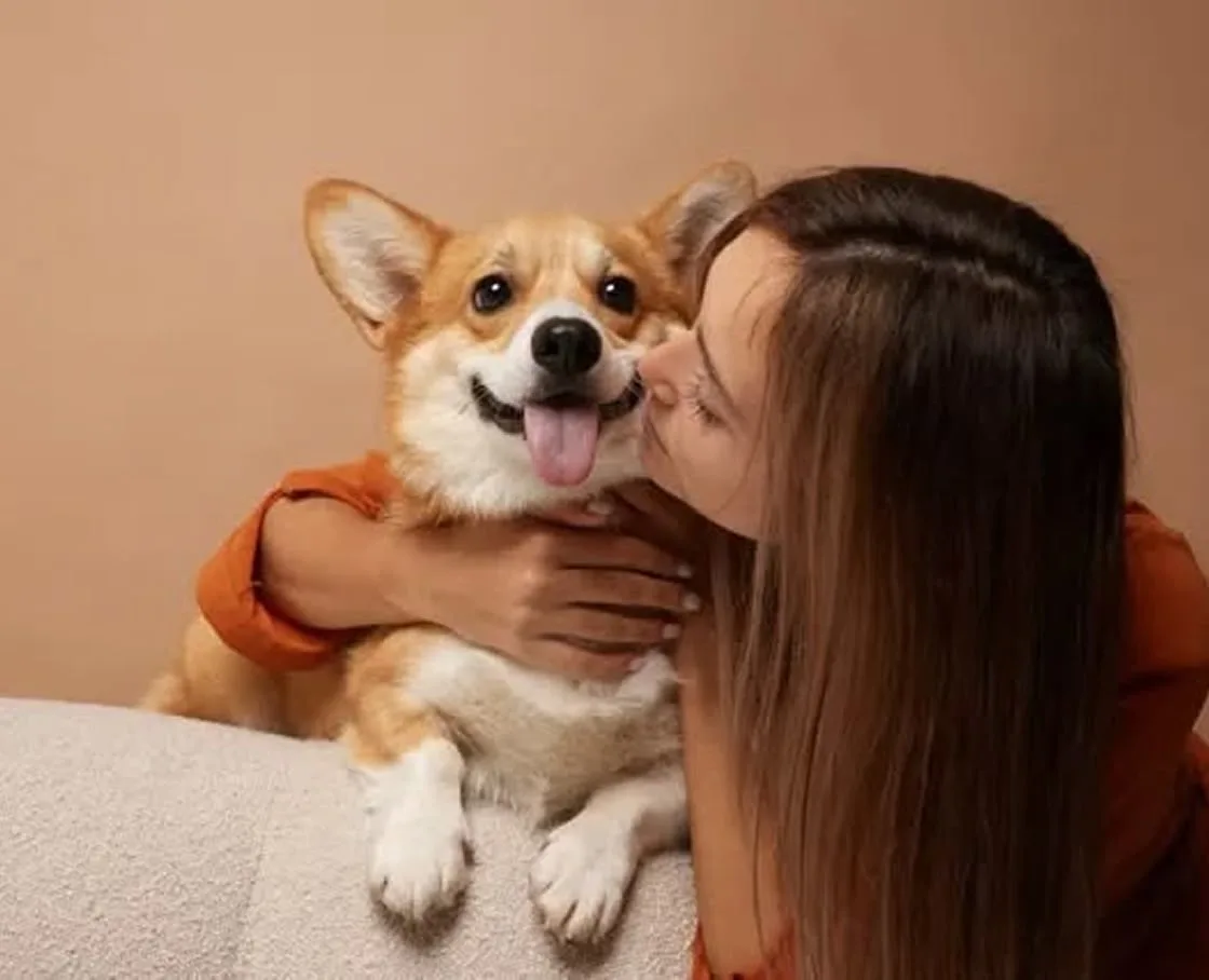 A woman with long brown hair leaning in to kiss a Pembroke Welsh Corgi on the cheek; the Corgi is smiling with its tongue out, and both are positioned against a warm, tan background.