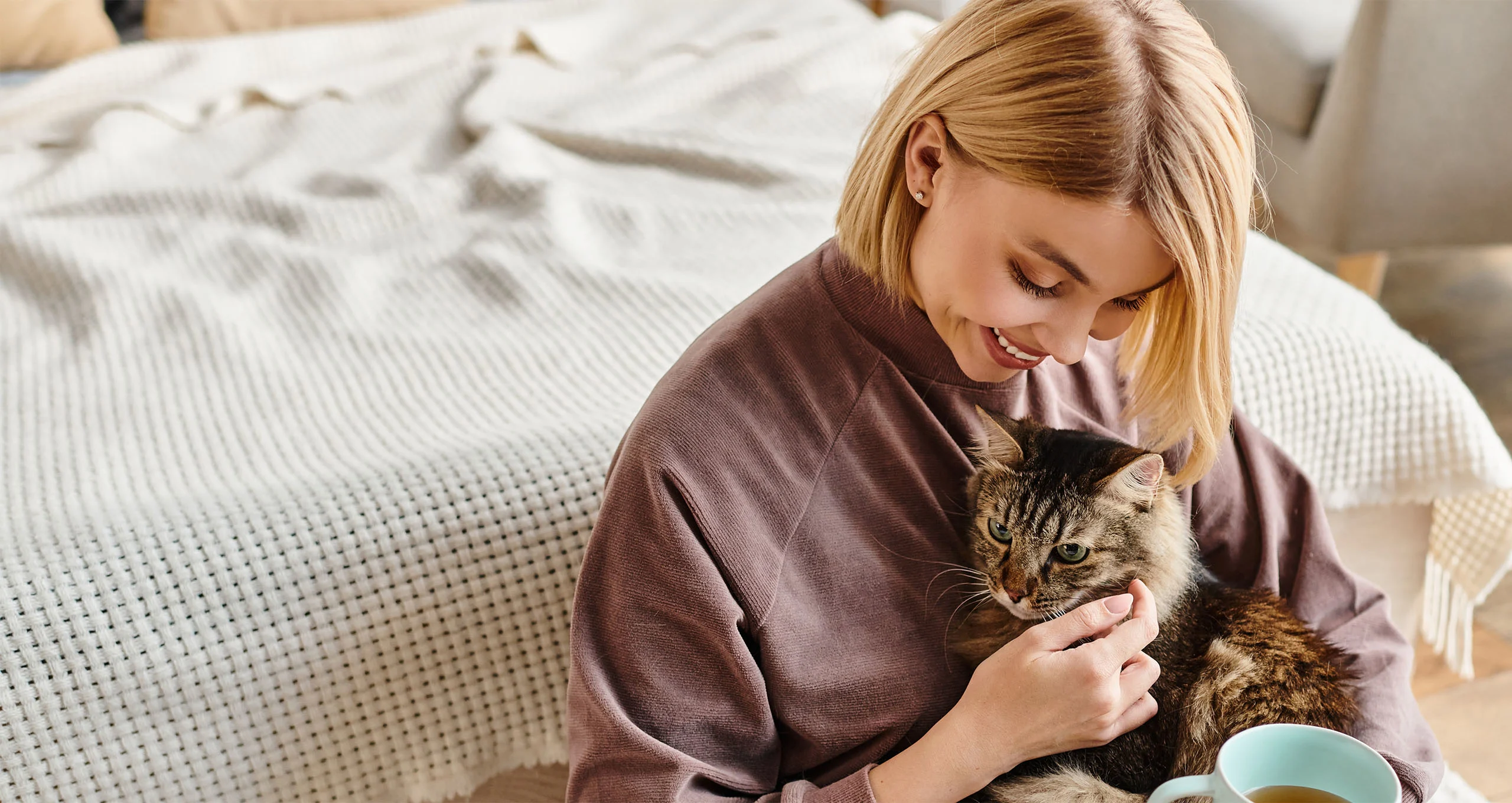 h woman in brown shirt hugging cat with cup of coffee in front of bed