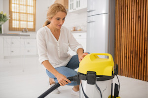 Woman in the kitchen about to vacuum behind and under the refrigerator and clean the refrigerator condenser coils.