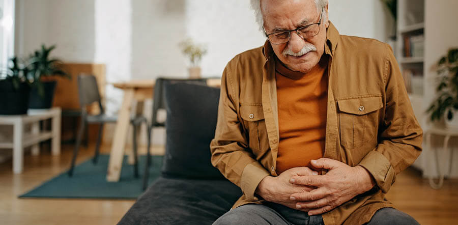 imagen que muestra a un señor de mediana edad sentado en un sofá apretándose con las manos en la zona del abdomen y con cara de dolor 