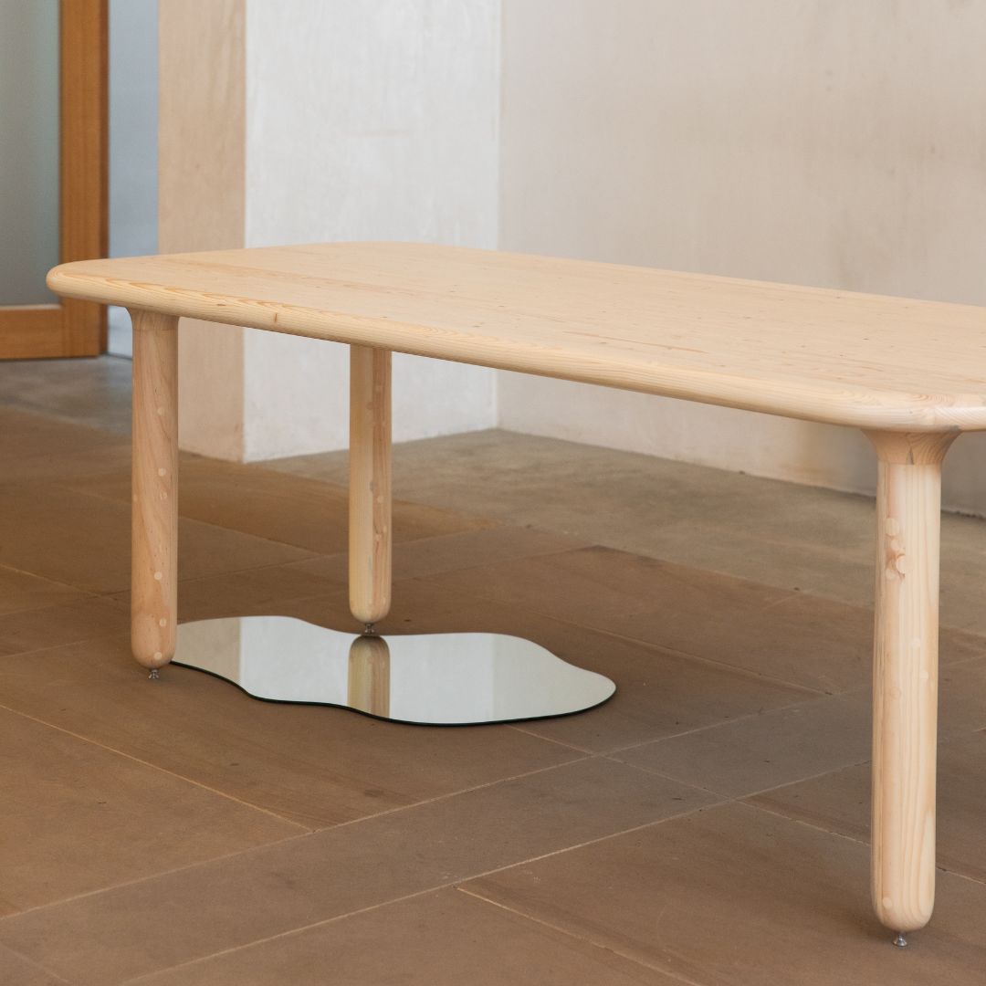 A pale wood dining table, smooth and rounded edges and legs. A puddle like mirror sits underneath to illustrate the way the legs join to the table top