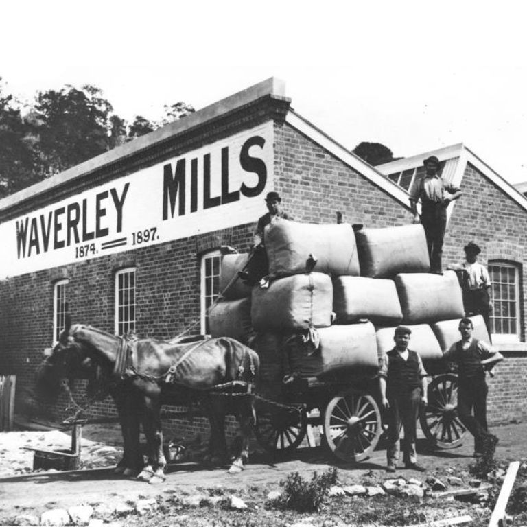 A team of horses are hitched to a wagon stacked with huge bales of wool in front of a Waverley Mills brick building 