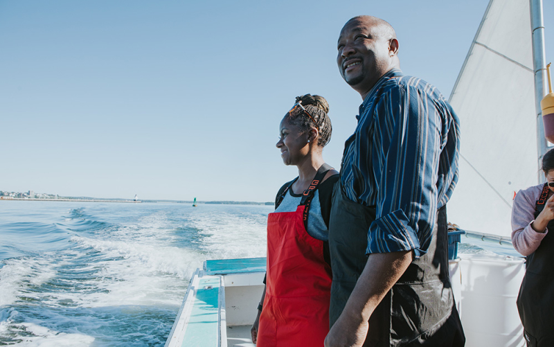 A photo of our two Memphis & OKC owners standing on a lobster boat in fishing gear, admiring the view of the coast of Maine.