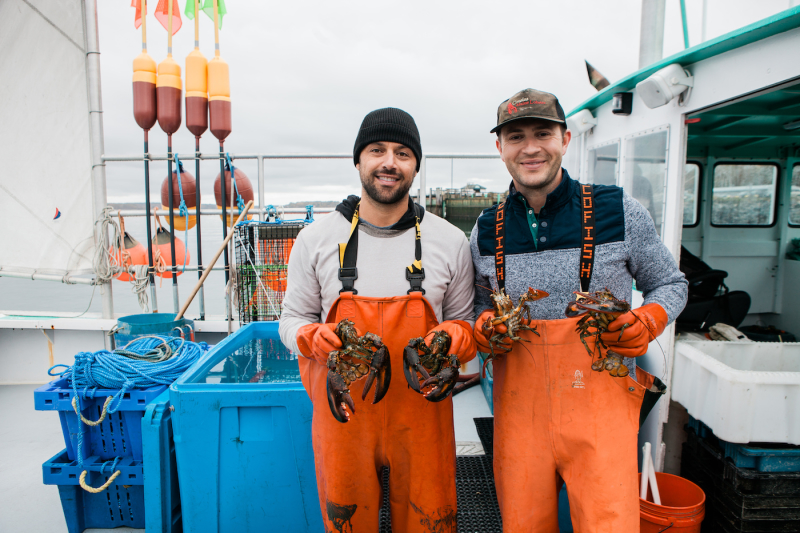 A photo of Cousins Maine Lobster founders, Jim and Sabin, on a fishing boat, dressed in fishing gear, holding live lobsters.