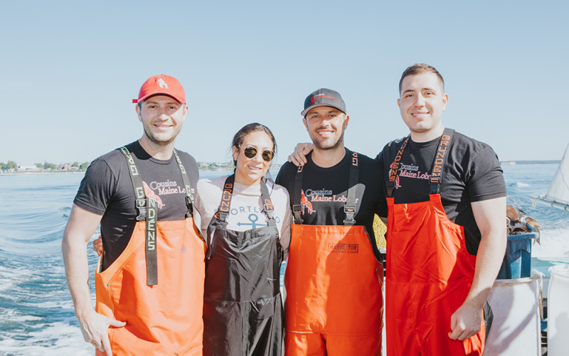 A photo of our Trenton & Philly franchisees standing on the back of a lobster boat in Maine with Cousins Maine Lobster founders, Jim & Sabin, all dressed in fishing gear.