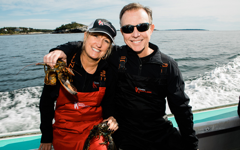 A photo of our Raleigh owners sitting on a gunwale of a lobster boat in Maine, holding live lobsters.