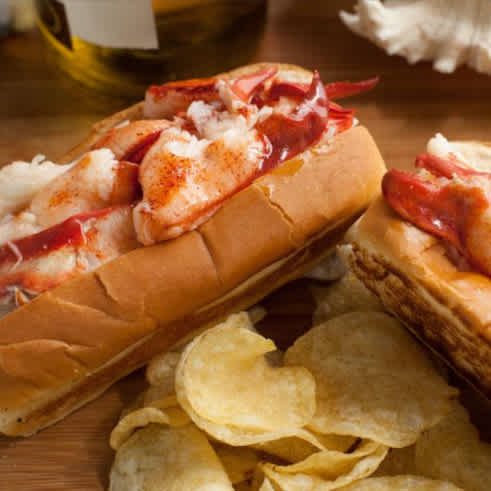 A photo of two lobsters rolls on a wood cutting board with some potato chips nearby.