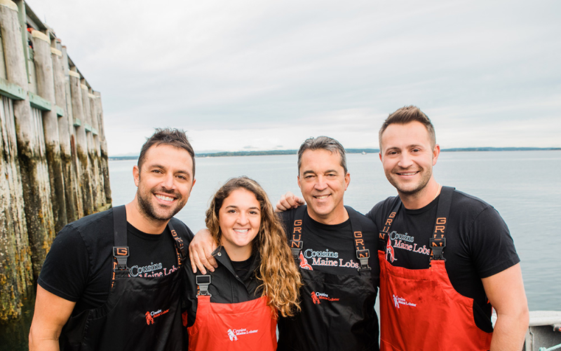 A photo of two members of our Boston family standing on the back of a fishing boat with Cousins Maine Lobster founders, Jim & Sabin, all dressed in fishing gear.