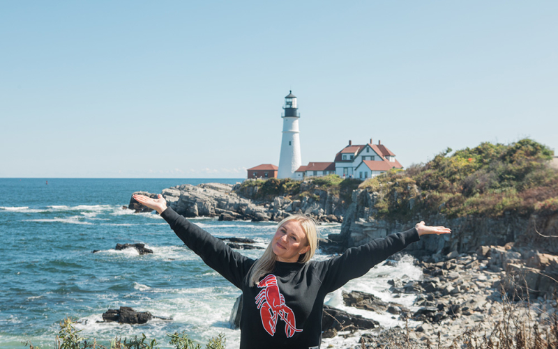A photo of our Freehold franchisee wearing a CML sweatshirt, standing in front of the Portland Head Light with her arms spread wide showcasing the lighthouse.