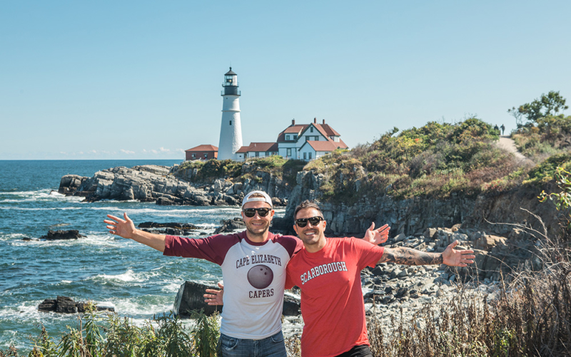 A photo of Cousins Maine Lobster founders, Jim & Sabin, standing in front of the Portland Head Lighthouse in Cape Elizabeth, Maine.
