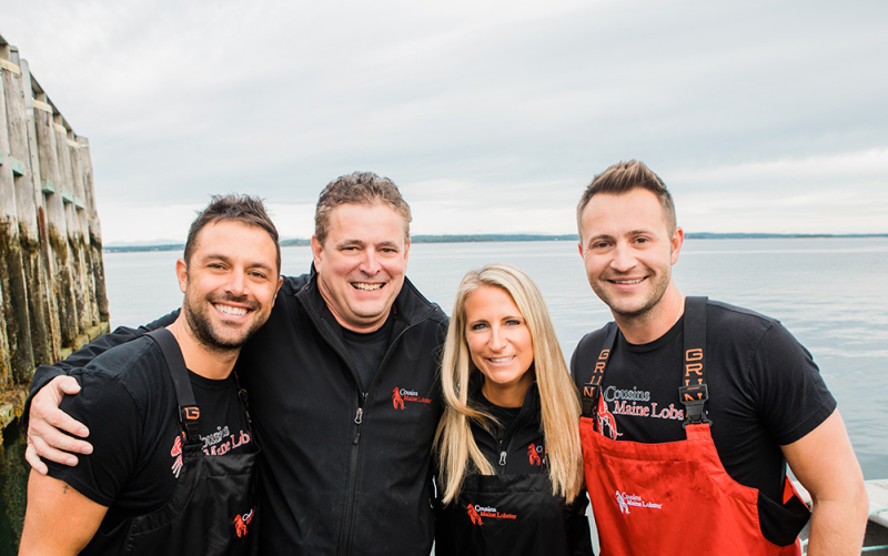 A photo of our Charlotte franchisees standing in Maine with Cousins Maine Lobster founders Jim & Sabin, all dressed in fishing gear.