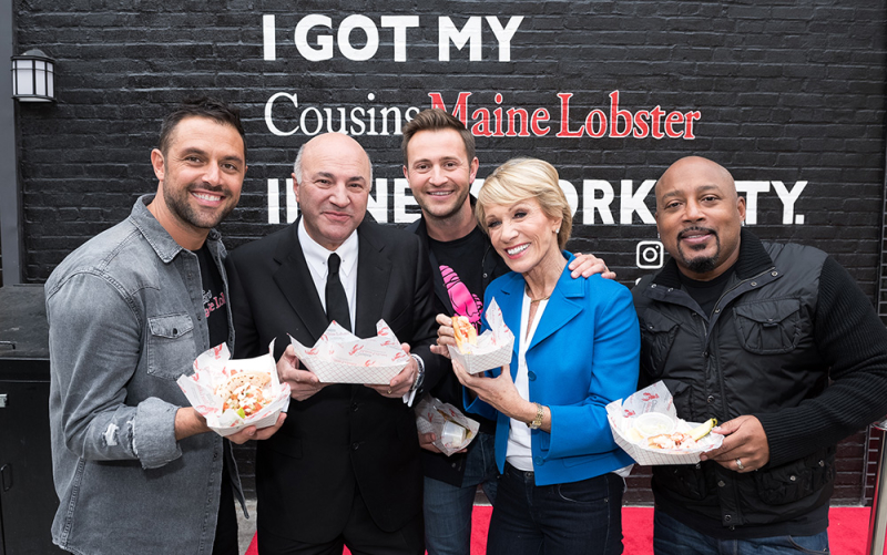 A photo of Jim & Sabin, in New York city, with Shark Tank sharks, Barbara, Daymond, and Mr. Wonderful, all holding Cousins Maine Lobster food items.
