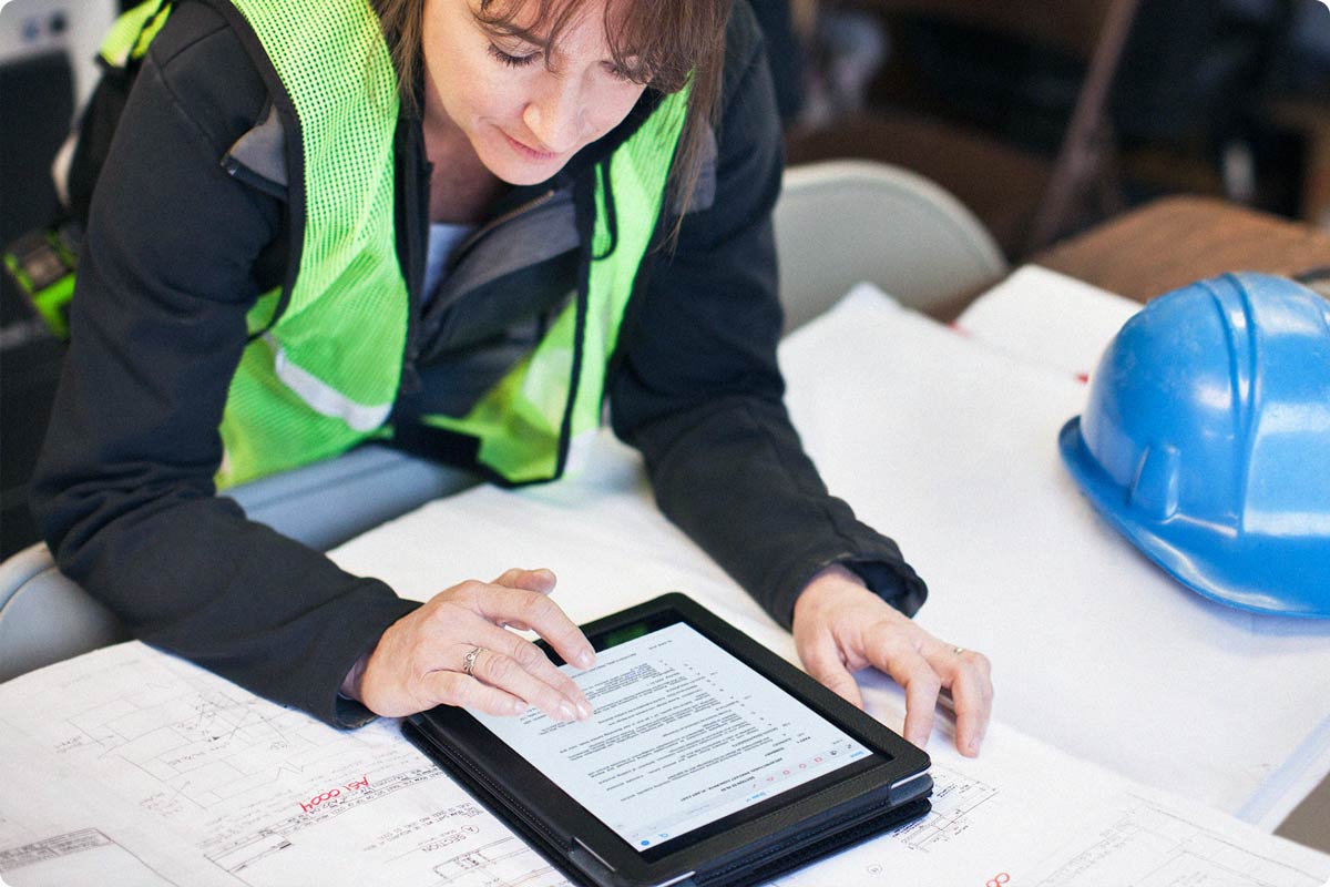 A female construction worker is in the jobsite trailer looking at their iPad. On the iPad she is viewing the Specifications for the project.