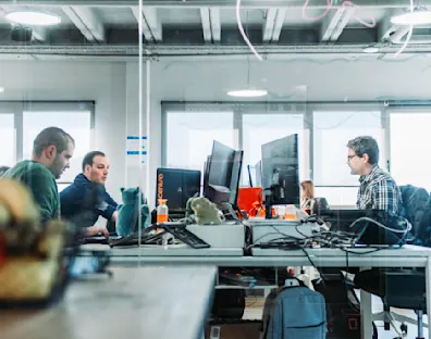 Imagen de una oficina con 3 personas detrás de un cristal en una oficina mirando sus monitores.