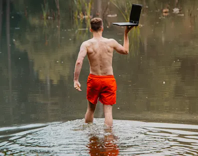 Imagen de un hombre en bañador metiéndose en un lago con un portátil en la mano derecha que lleva como si se tratase de una bandeja de bebidas