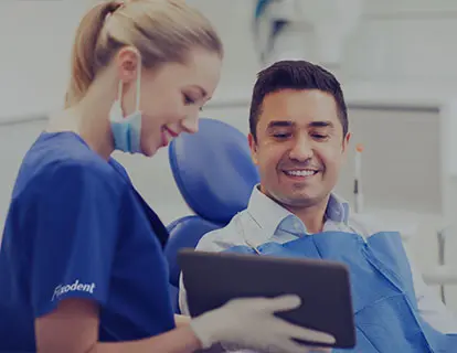 Une dentiste en blouse bleue tient en main une tablette qu'elle montre à son patient assis dans un fauteuil de dentiste. Il sourit car il a désormais les réponses à ses questions concernant les prothèses dentaires et les crèmes adhésives.