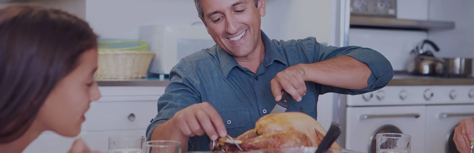 Un homme d'une cinquantaine d'années est assis à table en famille. Il est en train de découper un poulet. Il sourit car il sait comment profiter pleinement de sa crème adhésive pour prothèse dentaire.