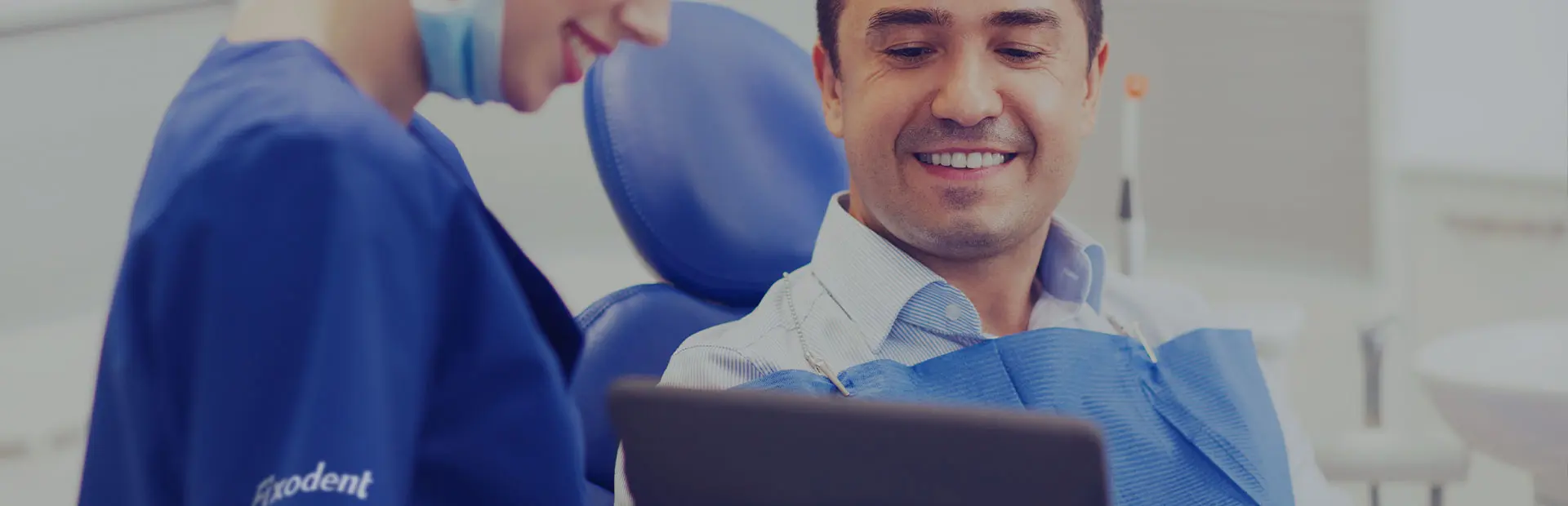 Une dentiste en blouse bleue tient en main une tablette qu'elle montre à son patient assis dans un fauteuil de dentiste. Il sourit car il a désormais les réponses à ses questions concernant les prothèses dentaires et les crèmes adhésives.