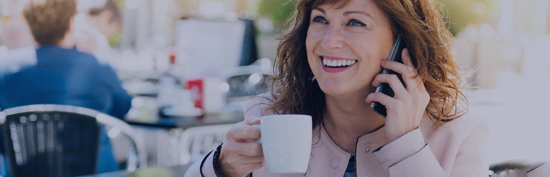 Une femme d'une cinquantaine d'années est assise à une terrasse et boit un café. Elle est au téléphone et sourit car elle sait qu'elle peut utiliser de la crème adhésive avec ses prothèses dentaires partielles.