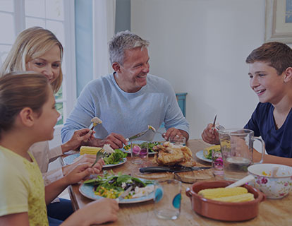 Une famille est assise autour de la table pour déjeuner. Toute la famille sourit y compris le père qui porte un dentier, car il connaît les avantages et inconvénients de porter sa prothèse dentaire avec ou sans crème adhésive.