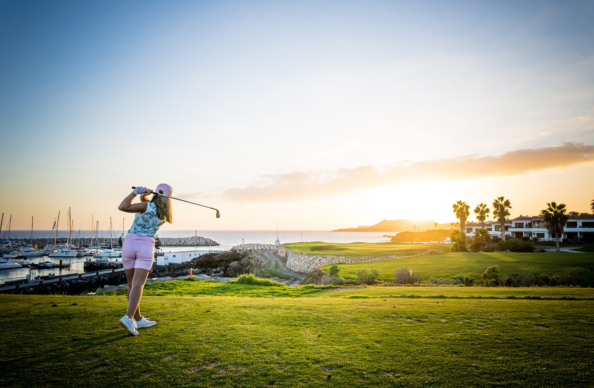 golfaria lyö avauslyöntiä rannikon golfkentällä. vihreät väylät johtavat kohti merta, taustalla satama ja rakennuksia lämpimässä iltavalossa ja matalan auringon aikaan.