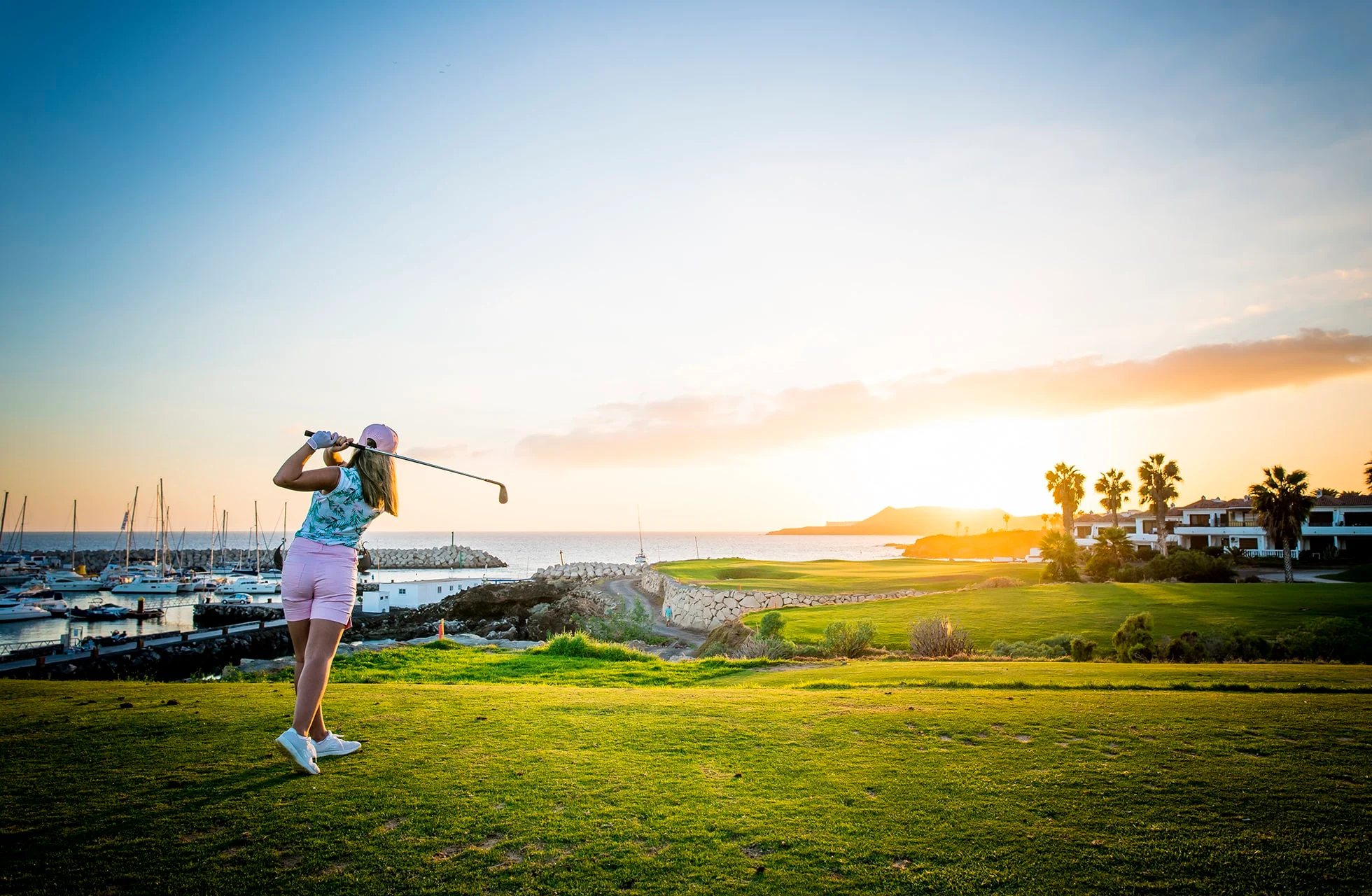 golfaria lyö avauslyöntiä rannikon golfkentällä. vihreät väylät johtavat kohti merta, taustalla satama ja rakennuksia lämpimässä iltavalossa ja matalan auringon aikaan.