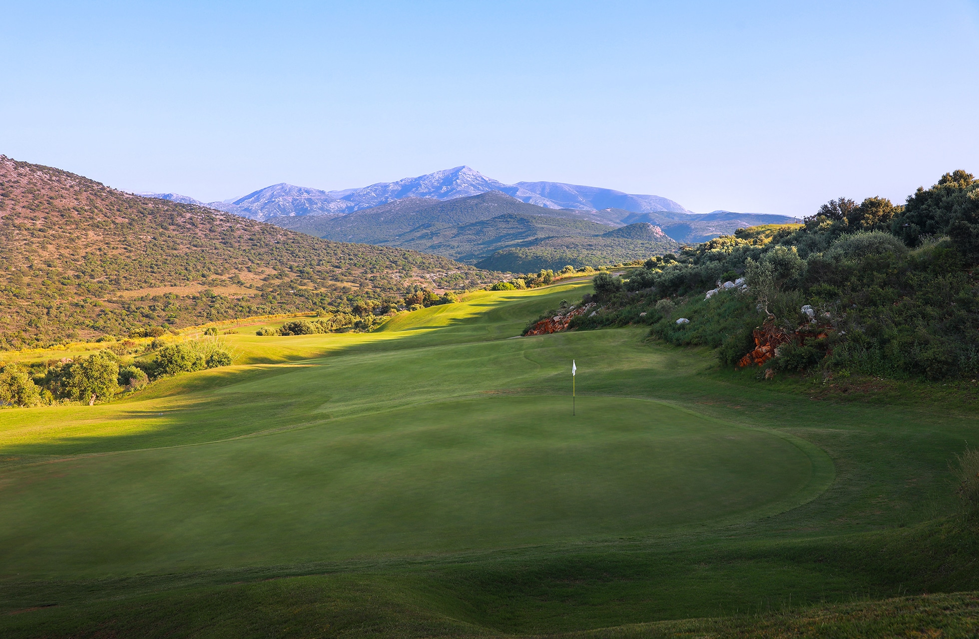 golfgreen med flagg omgitt av bølgende fairway, med åser og fjell i bakgrunnen i klart dagslys