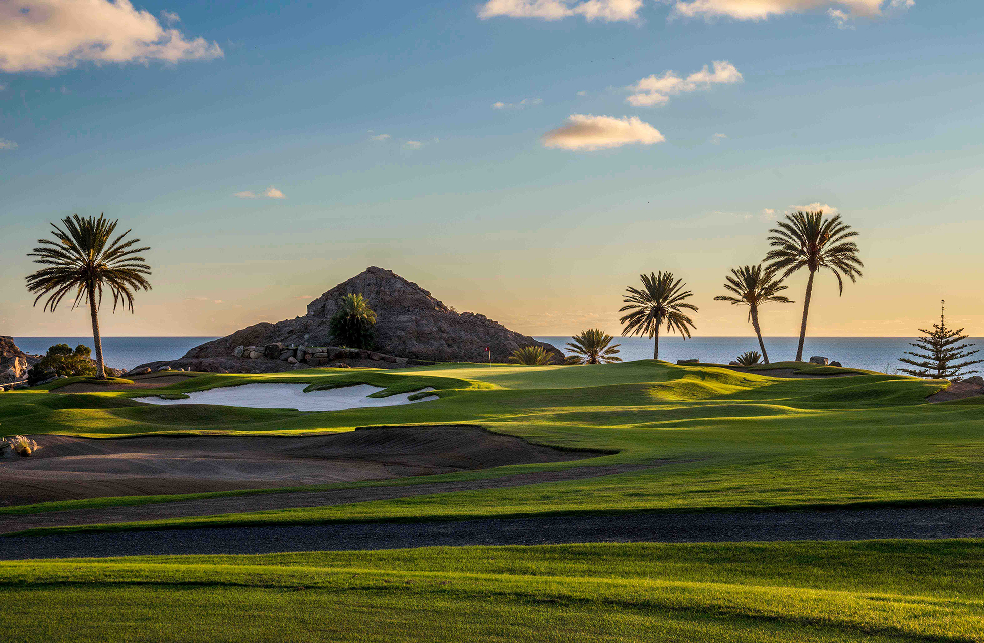 golfgreen på anfi tauro golf med bunkere, bølgende fairwayer og palmer i kupert landskap, med fjell og hav i bakgrunnen i varmt kveldslys