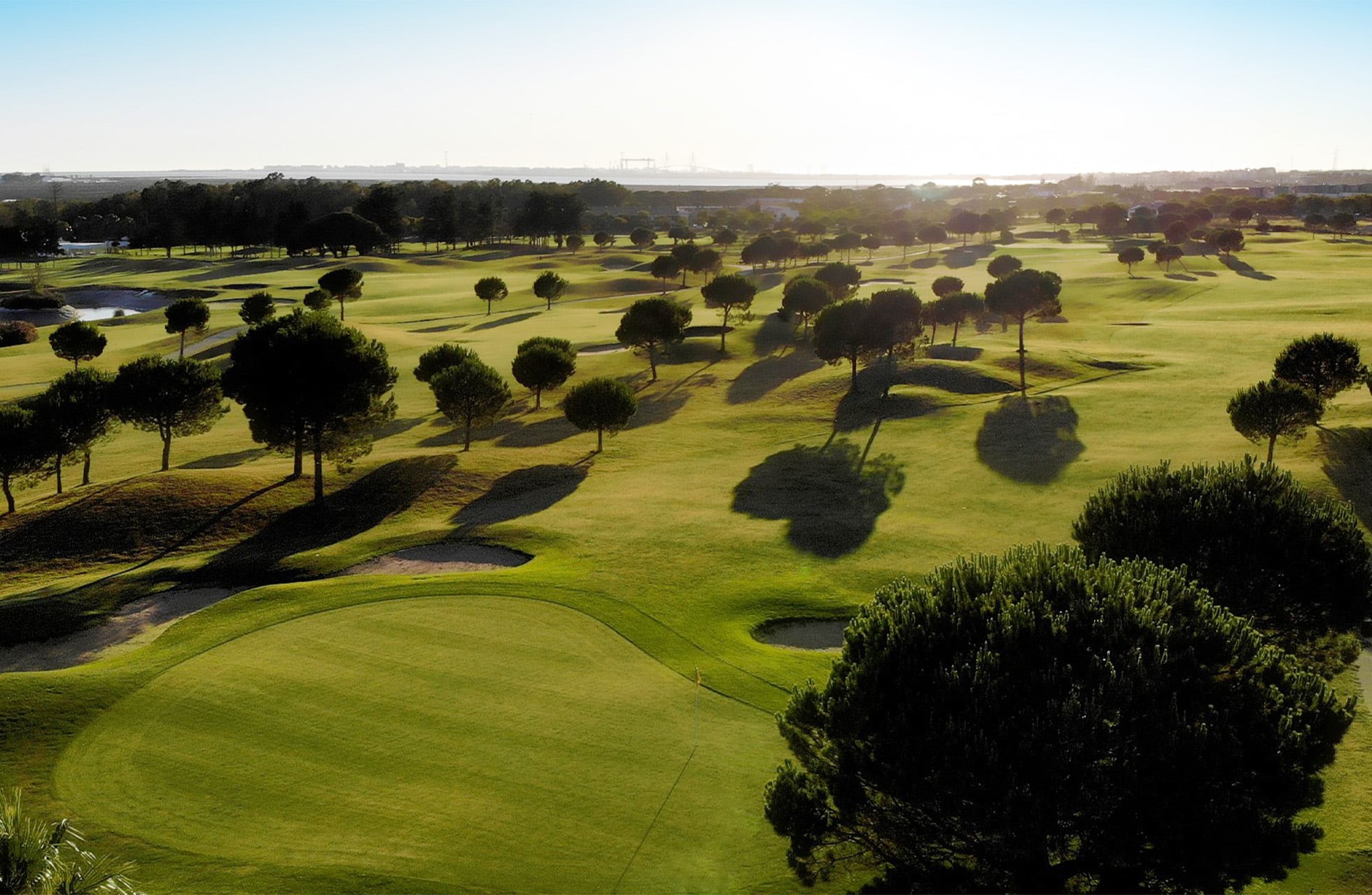Luftfoto av en solbelyst golfbane med trær, greener og bunkere