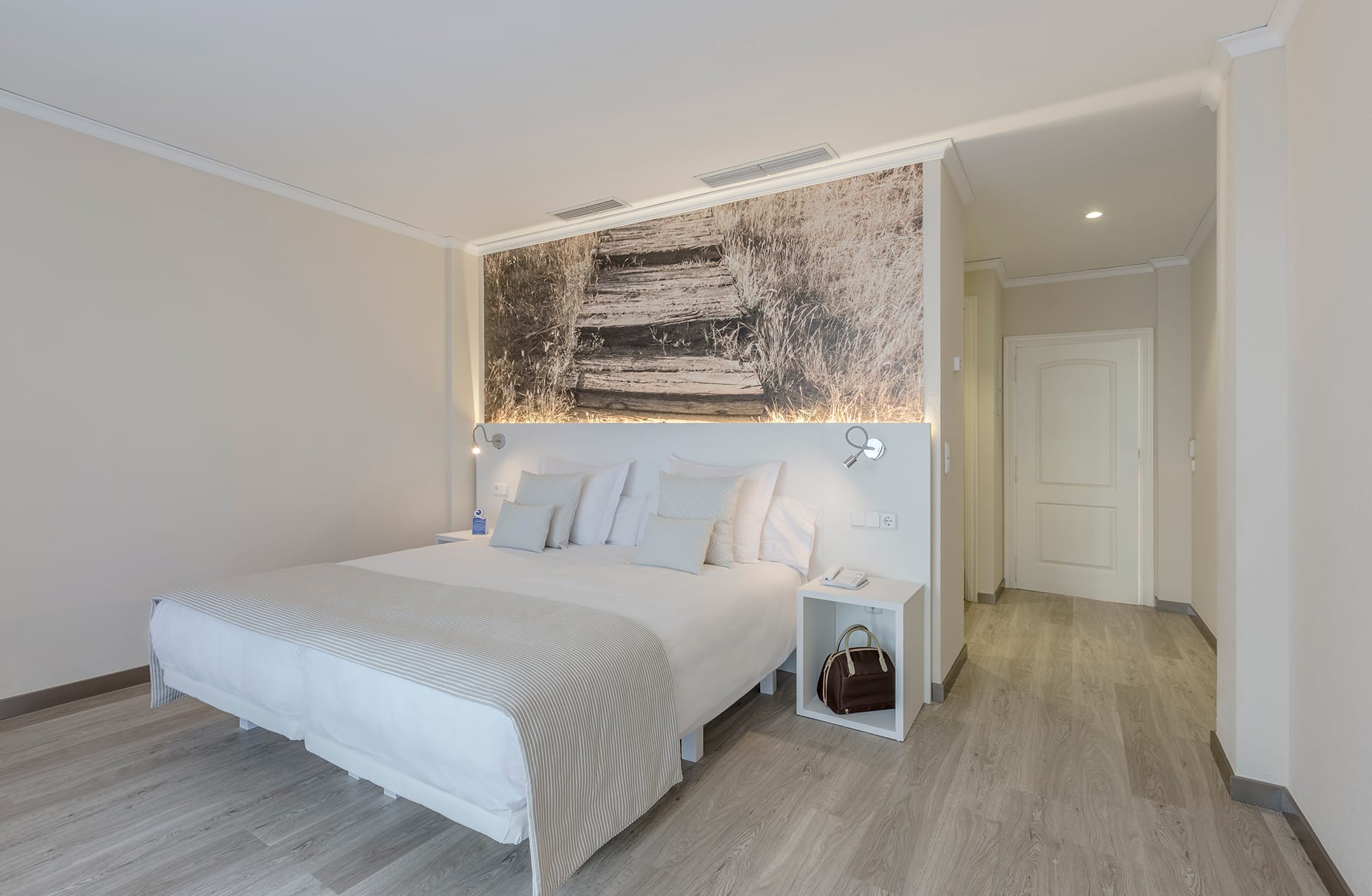lyst og moderne hotelværelse med dobbeltseng, sengeborde med lamper, dekorativ bagvæg med kunstværk samt lyse gulve og gang mod døren i dagslys