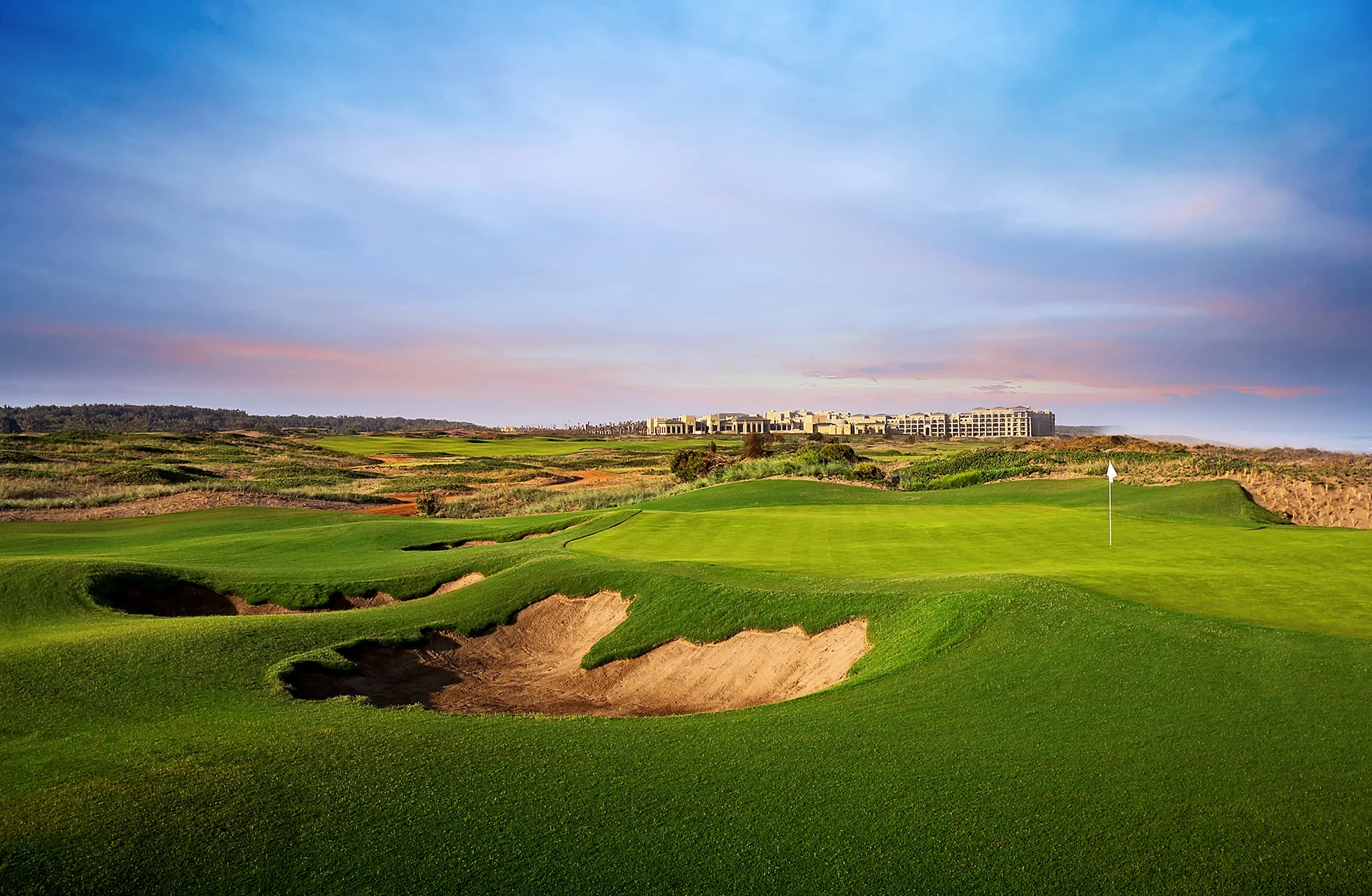 grønn golfbane med sandbunkere, flagg på greenen og bygninger i bakgrunnen i kystlandskap