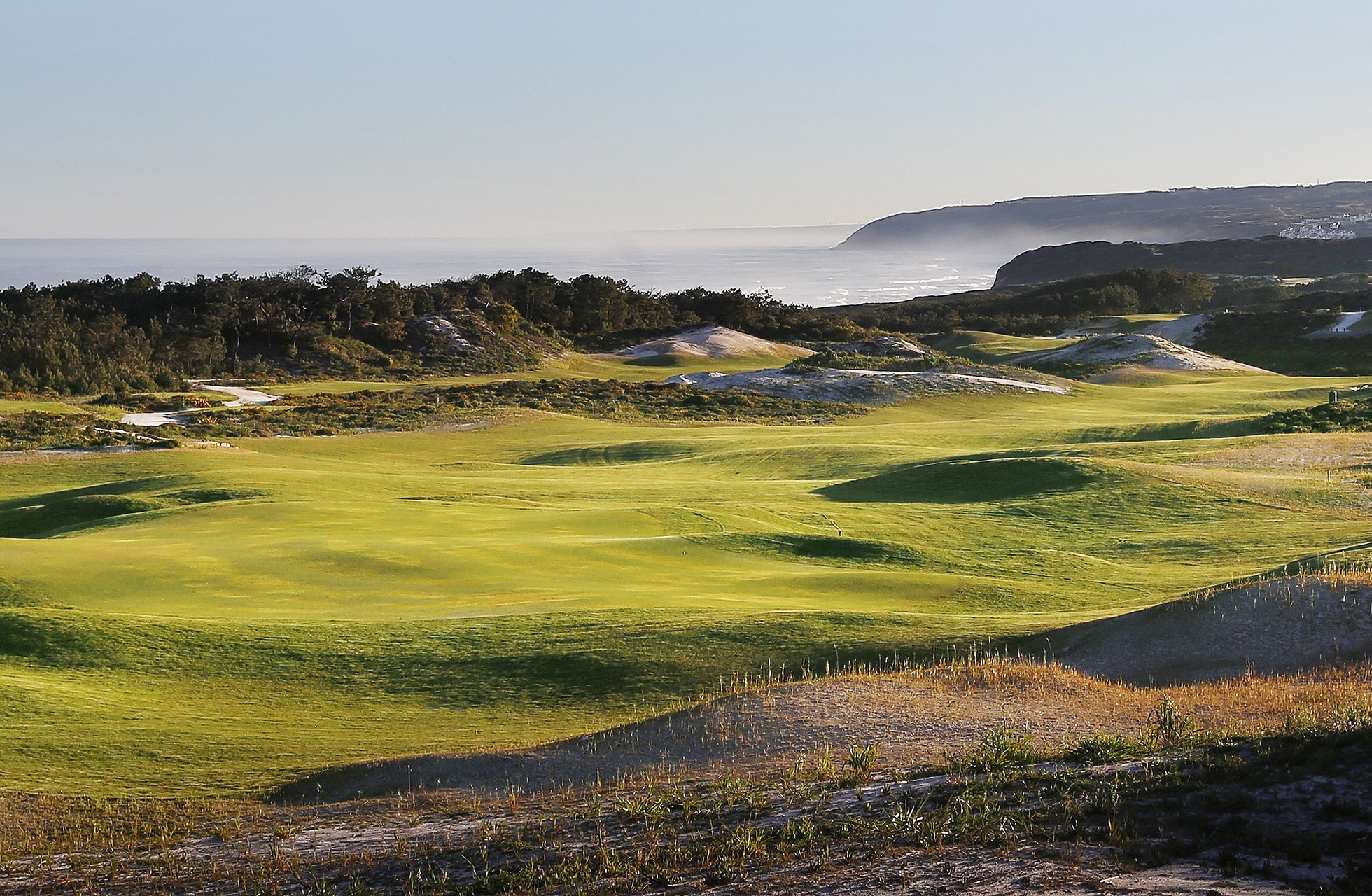 bølgende golfbane med mykt formede fairwayer og greener gjennom naturlig vegetasjon, med havet og kystlinjen synlig i bakgrunnen i klart dagslys