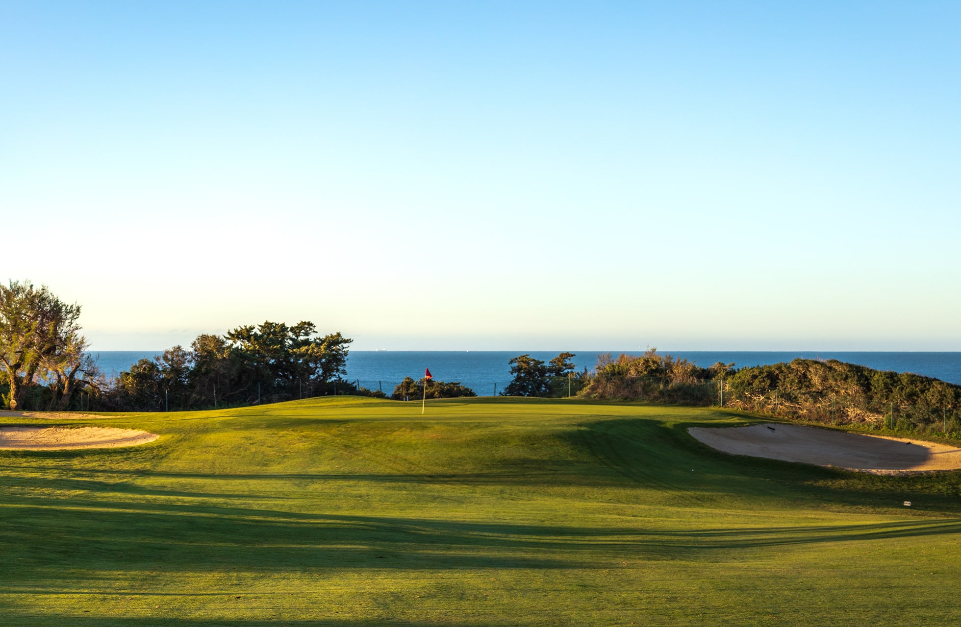 golfgreen med flagg og bunkere i åpent kystlandskap, med havet og horisonten i bakgrunnen i varmt kveldslys