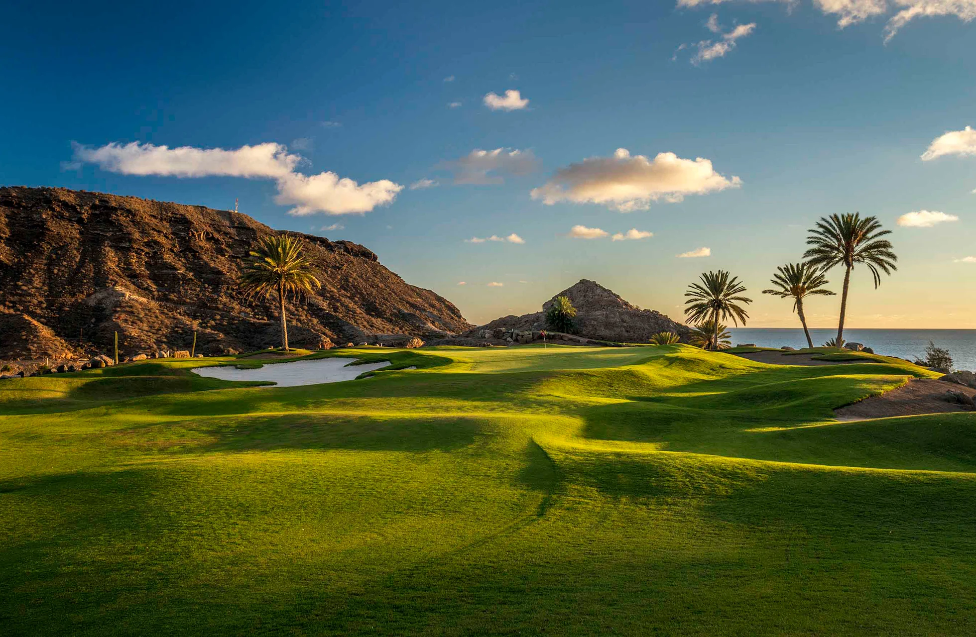 golfbane på anfi tauro golf med grønne fairwayer, bunkere, palmer og kupert, tørt landskap med fjell og hav i bakgrunnen i varmt kveldslys