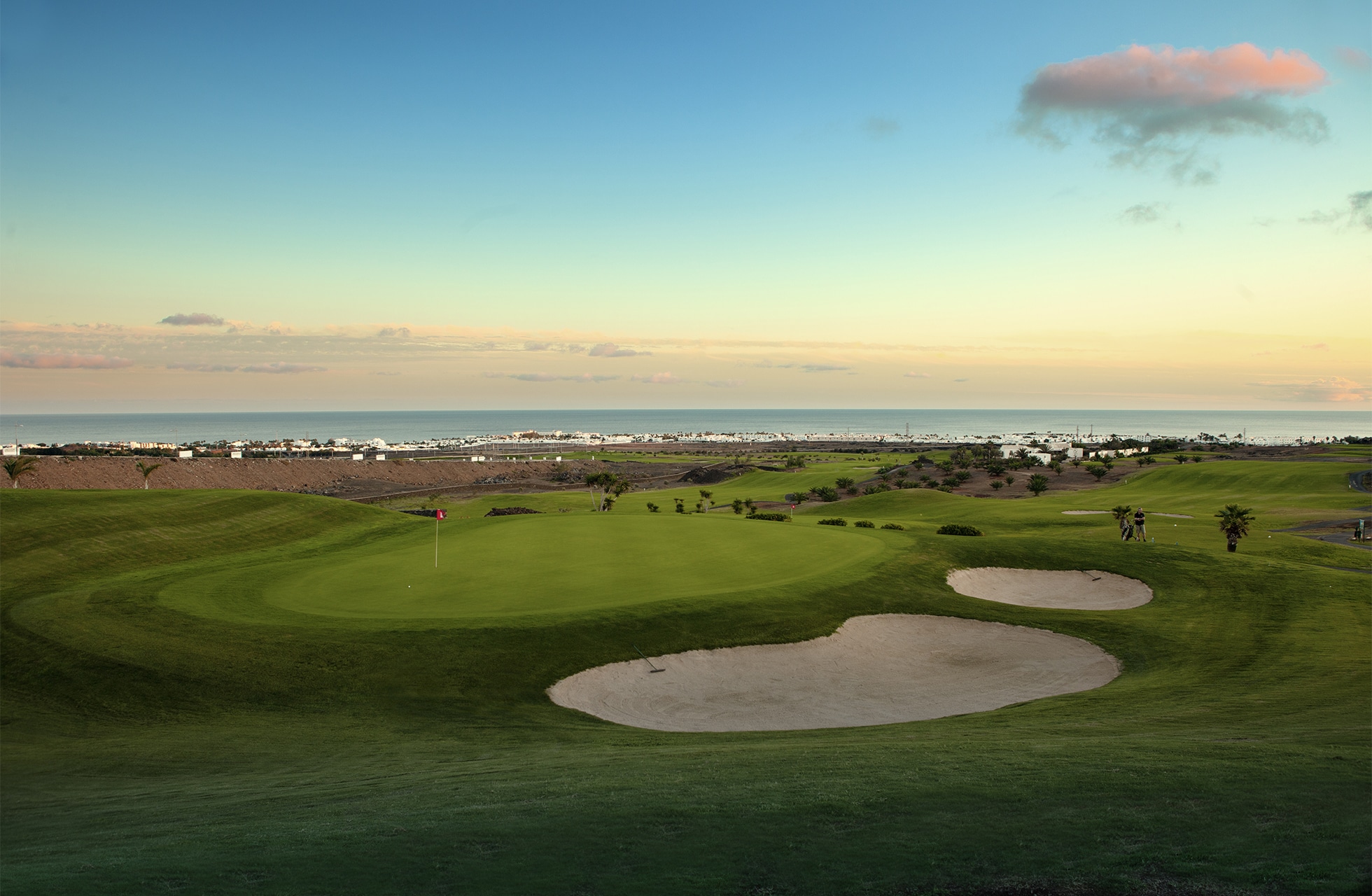 golfgreen med flagga och sandbunkrar i ett öppet landskap, med havet och kustbebyggelse i bakgrunden under mjukt kvällsljus.