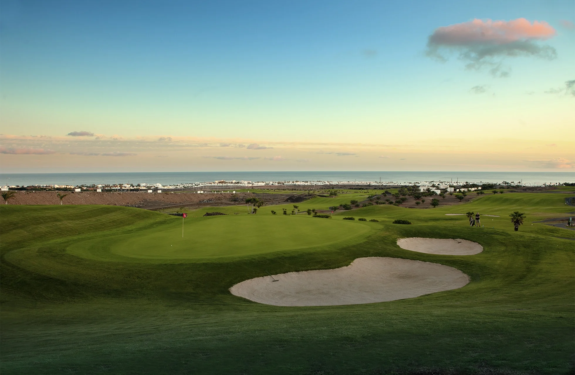 golfgreen med flagga och sandbunkrar i ett öppet landskap, med havet och kustbebyggelse i bakgrunden under mjukt kvällsljus.