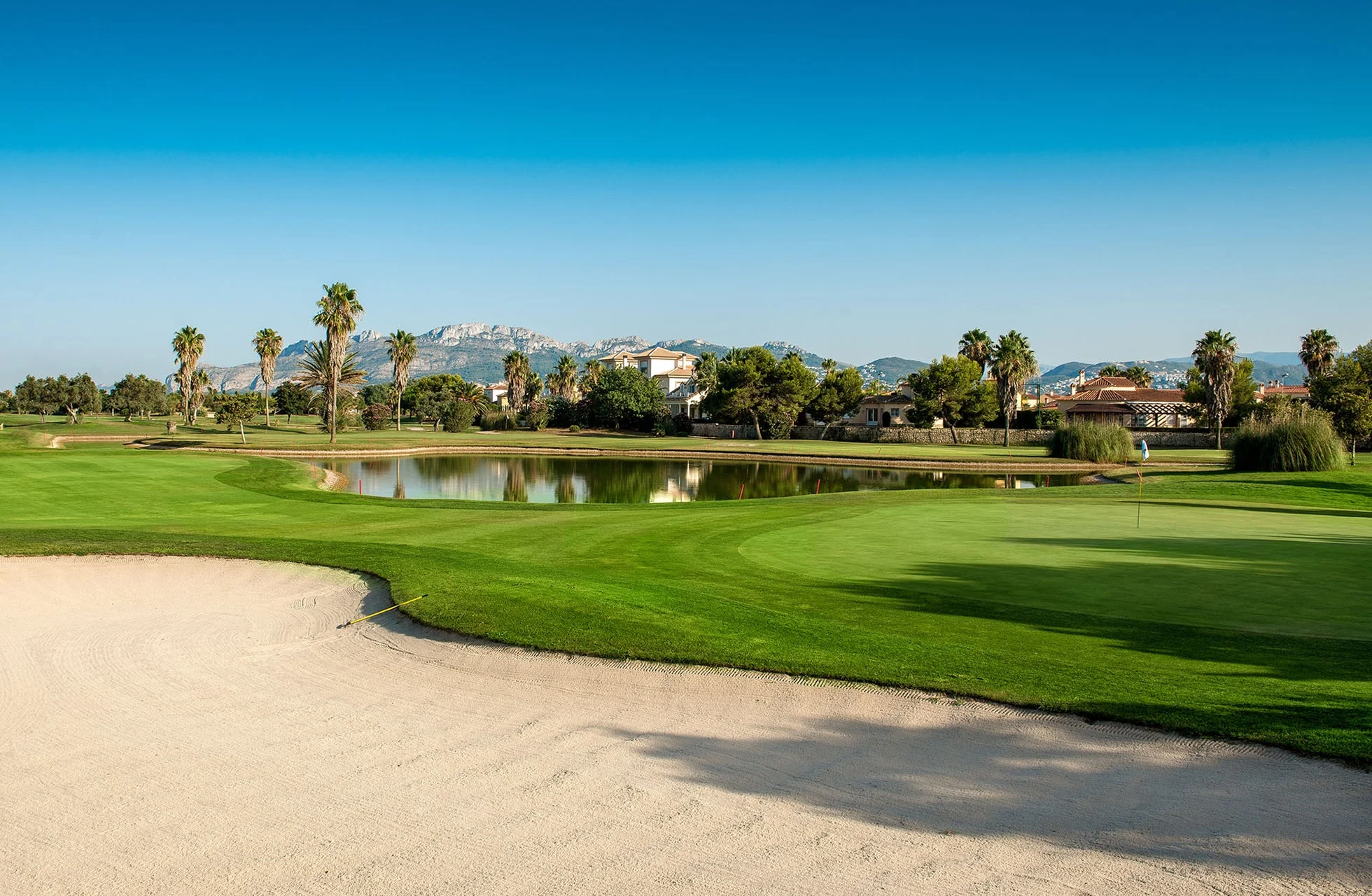 sandbunker i forgrunden, velplejet green og fairway langs et vandhazard, med palmer, bygninger og bjerge i baggrunden under klar blå himmel i dagslys