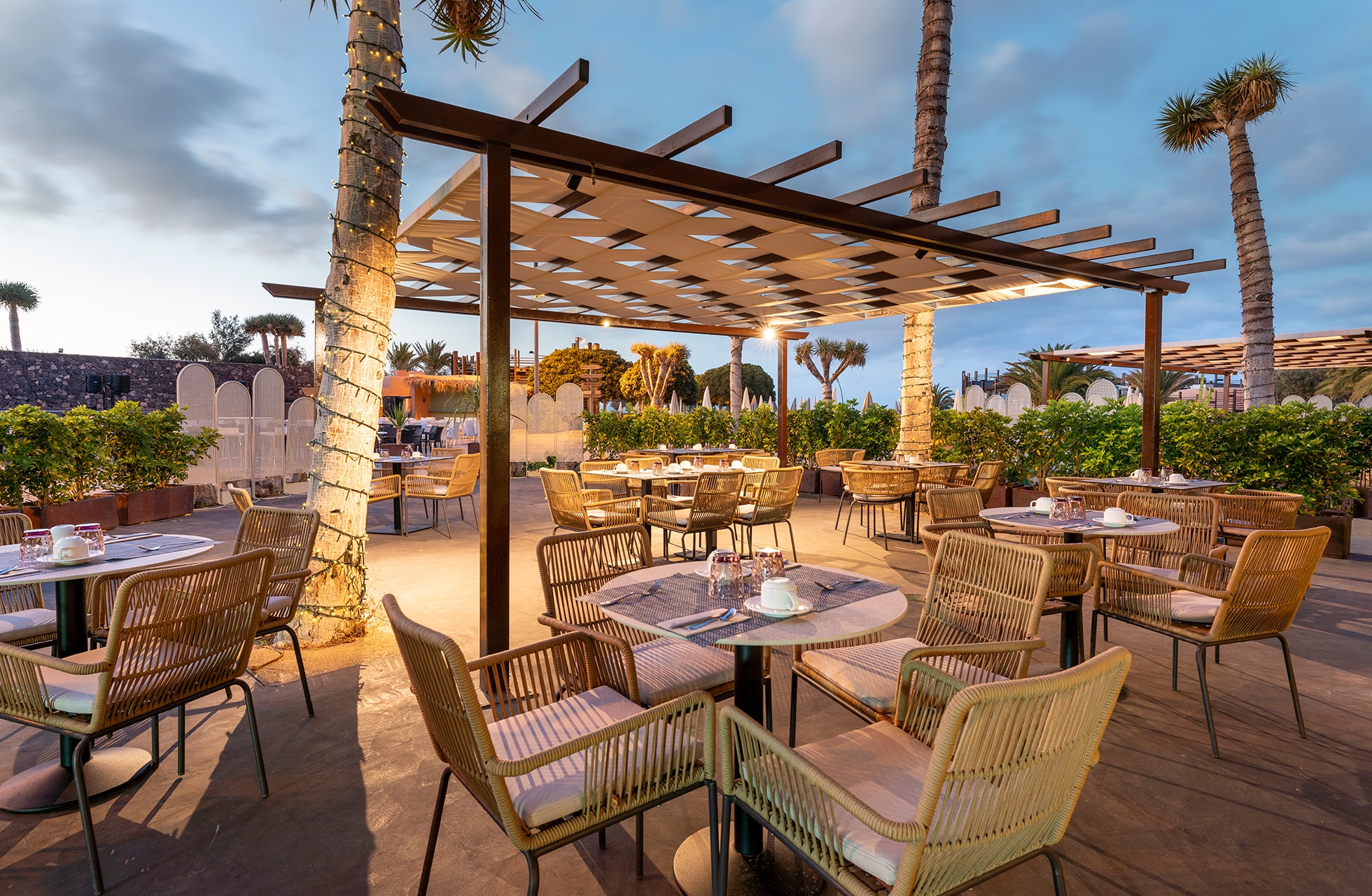 utendørs restaurant med dekkede bord og stoler under en pergola i tre. området er omgitt av palmer og grøntområder, med varm belysning og hotellbygninger i bakgrunnen i kveldslys.