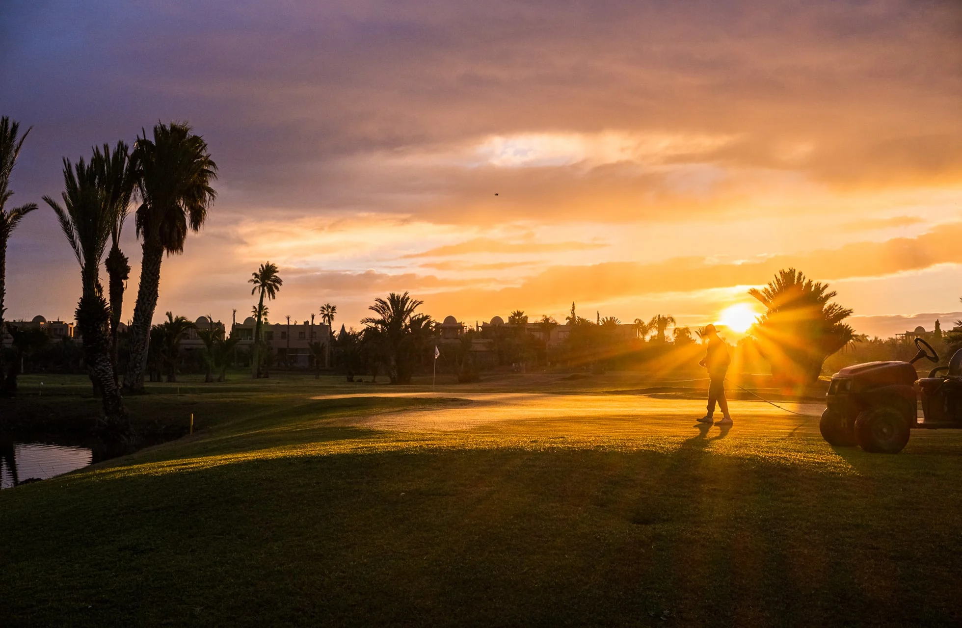 henkilö kävelee golfkentällä golfkärryn kanssa auringonlaskussa, palmuja, vesieste ja väylä lämpimässä iltavalossa