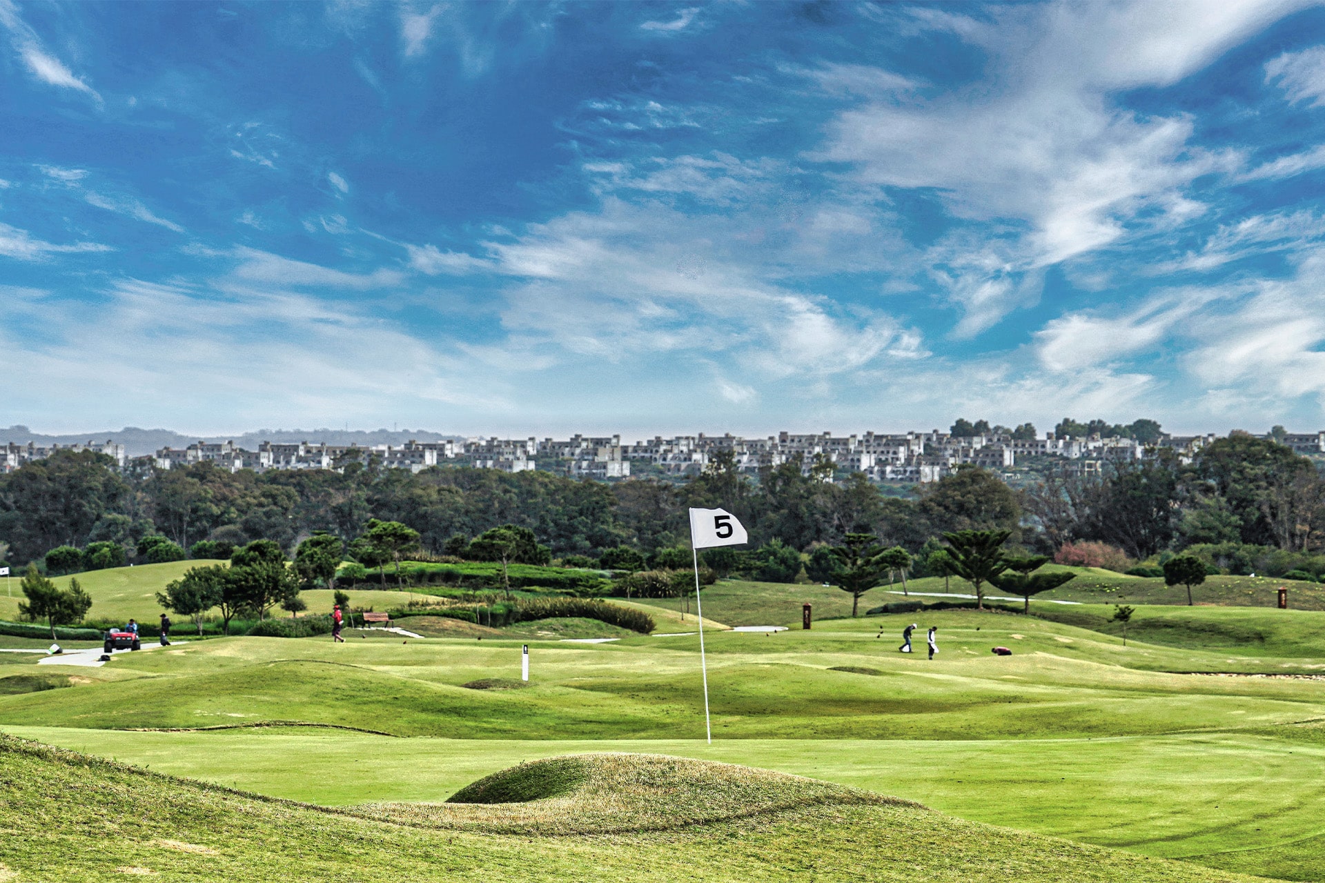 golfgreen med flag markeret med tallet 5, omgivet af bølgende fairways og træer, med bebyggelse synlig i baggrunden under klar blå himmel
