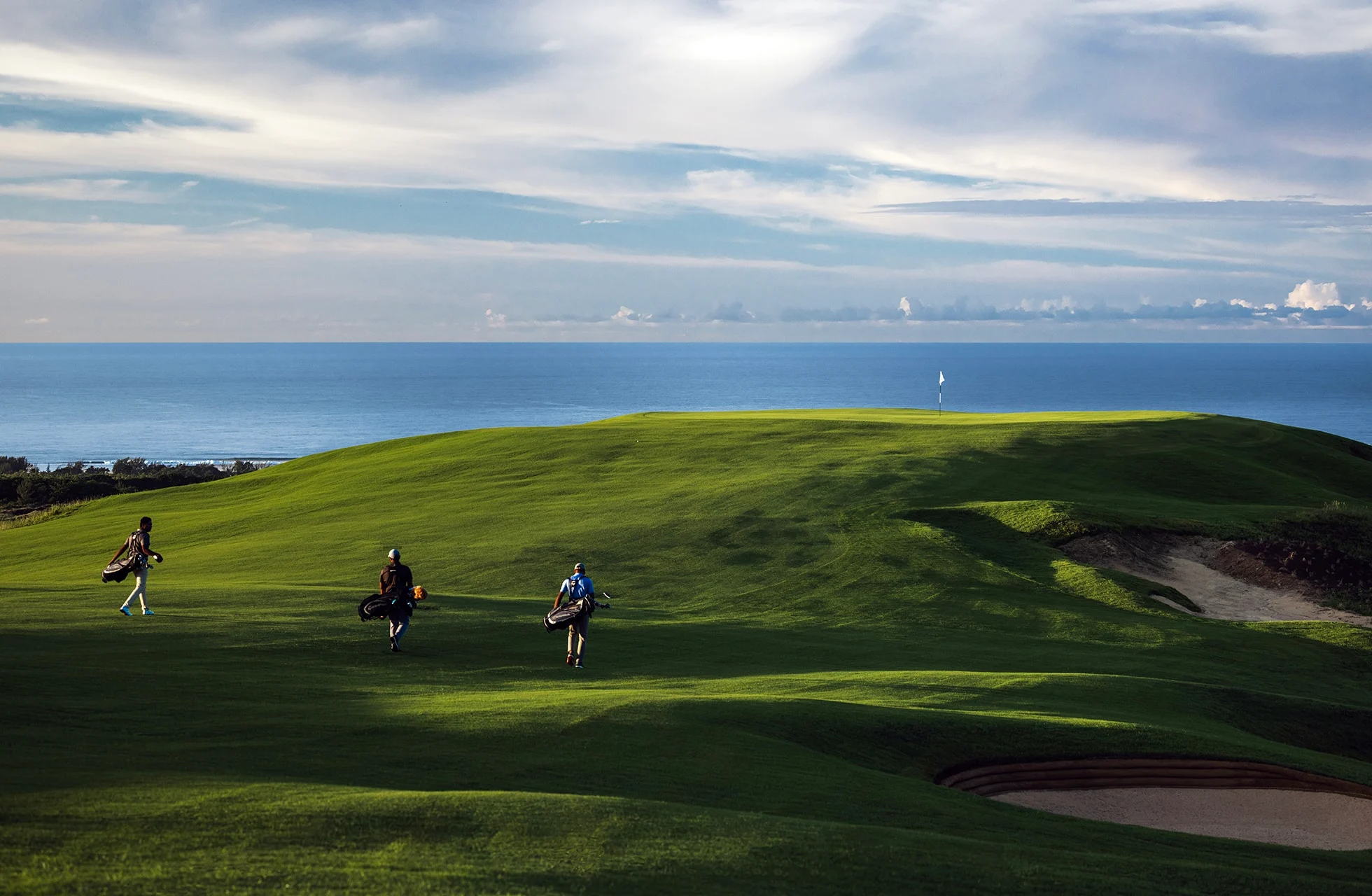 golfspillere går over bølgende green på heritage golf club la réserve golf links med bunker og åbent hav i baggrunden