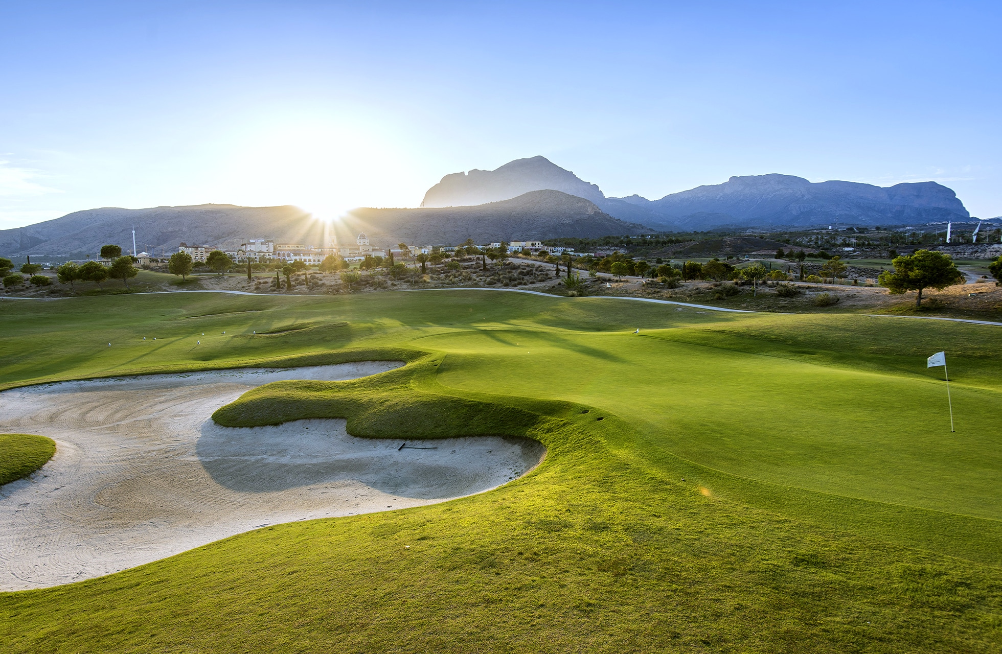 golfgreen med flere bunkere i forgrunnen og bølgende fairwayer gjennom landskapet, med fjell i bakgrunnen og solen lavt over horisonten i varmt kveldslys