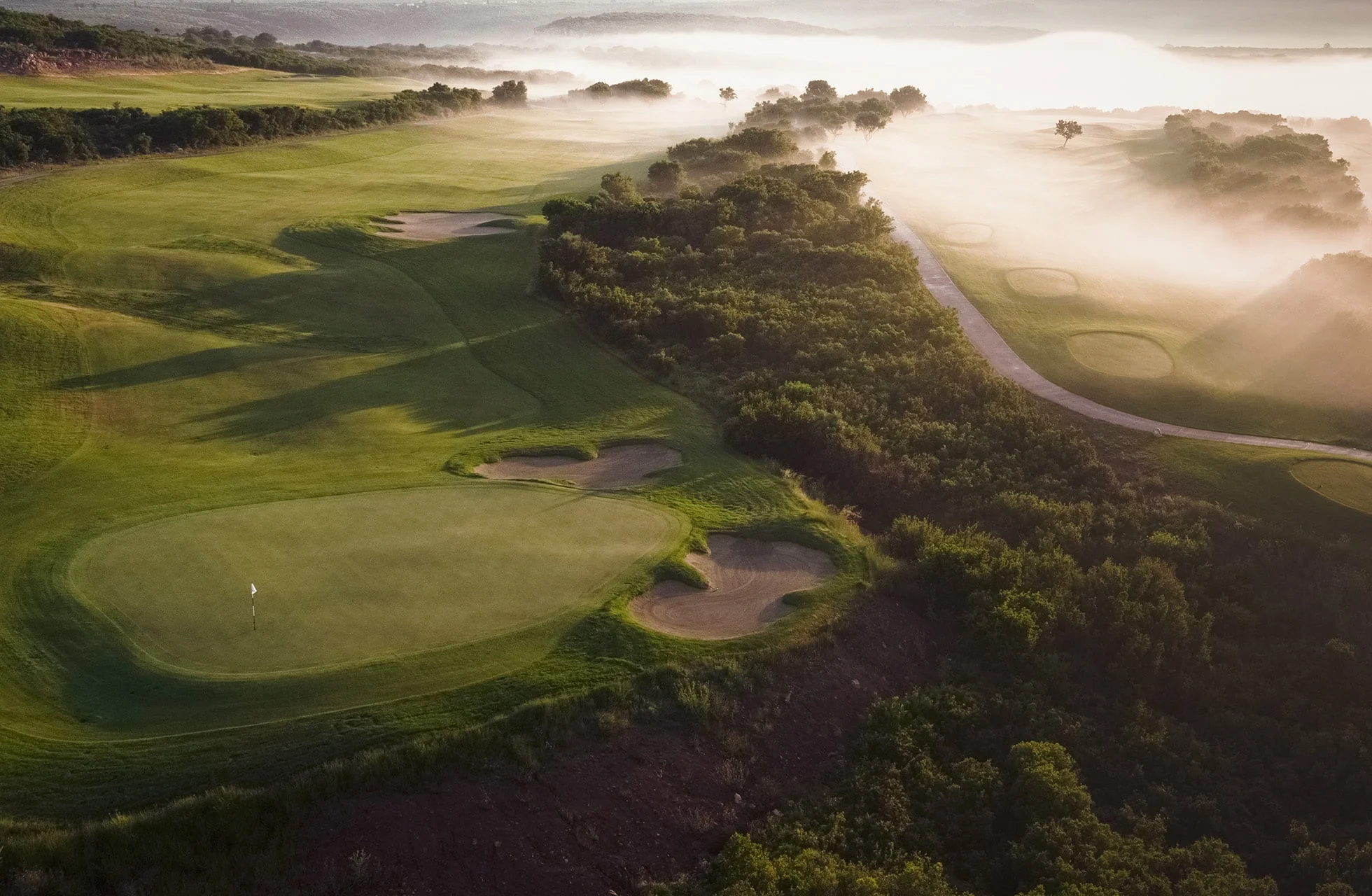 green med sandbunkere omgitt av tett vegetasjon og lett dis over golfbanen i morgenlys