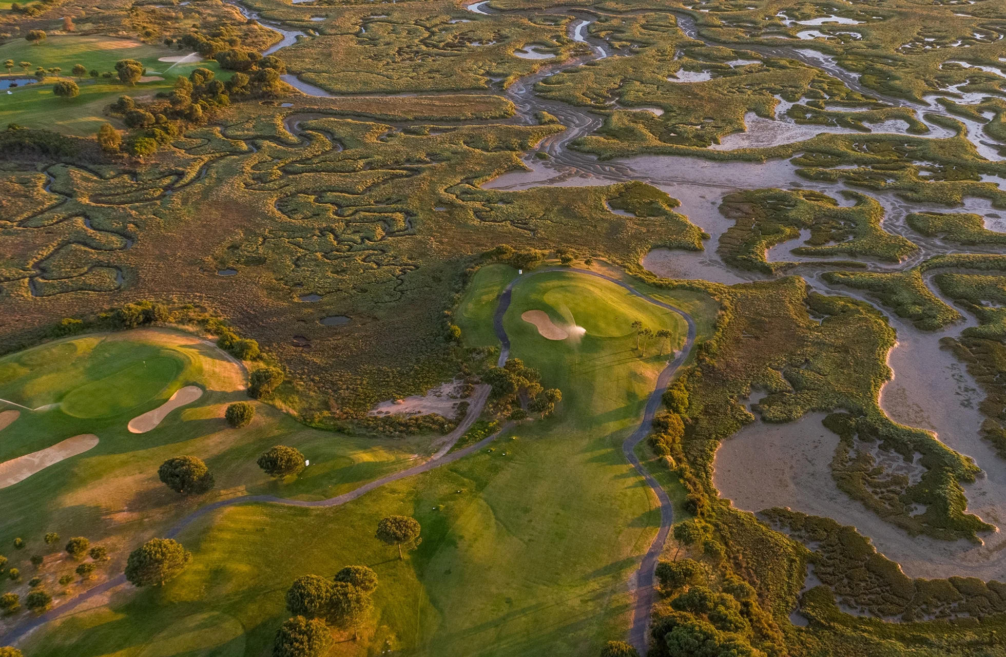flyfoto av en golfbane med green og fairway som slynger seg gjennom et vidstrakt våtmarksområde med kanaler, vegetasjon og naturlige vannflater i varmt kveldslys