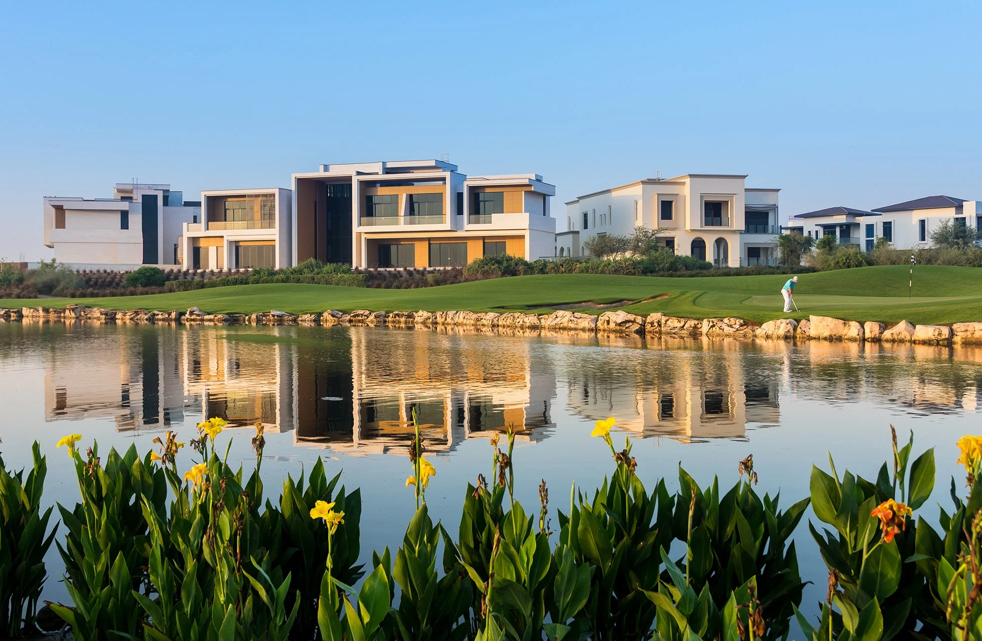 moderne villaer langs en golfbane med vann i forgrunnen som speiler bygningene, grønn fairway og en golfspiller synlig i bakgrunnen under klar himmel