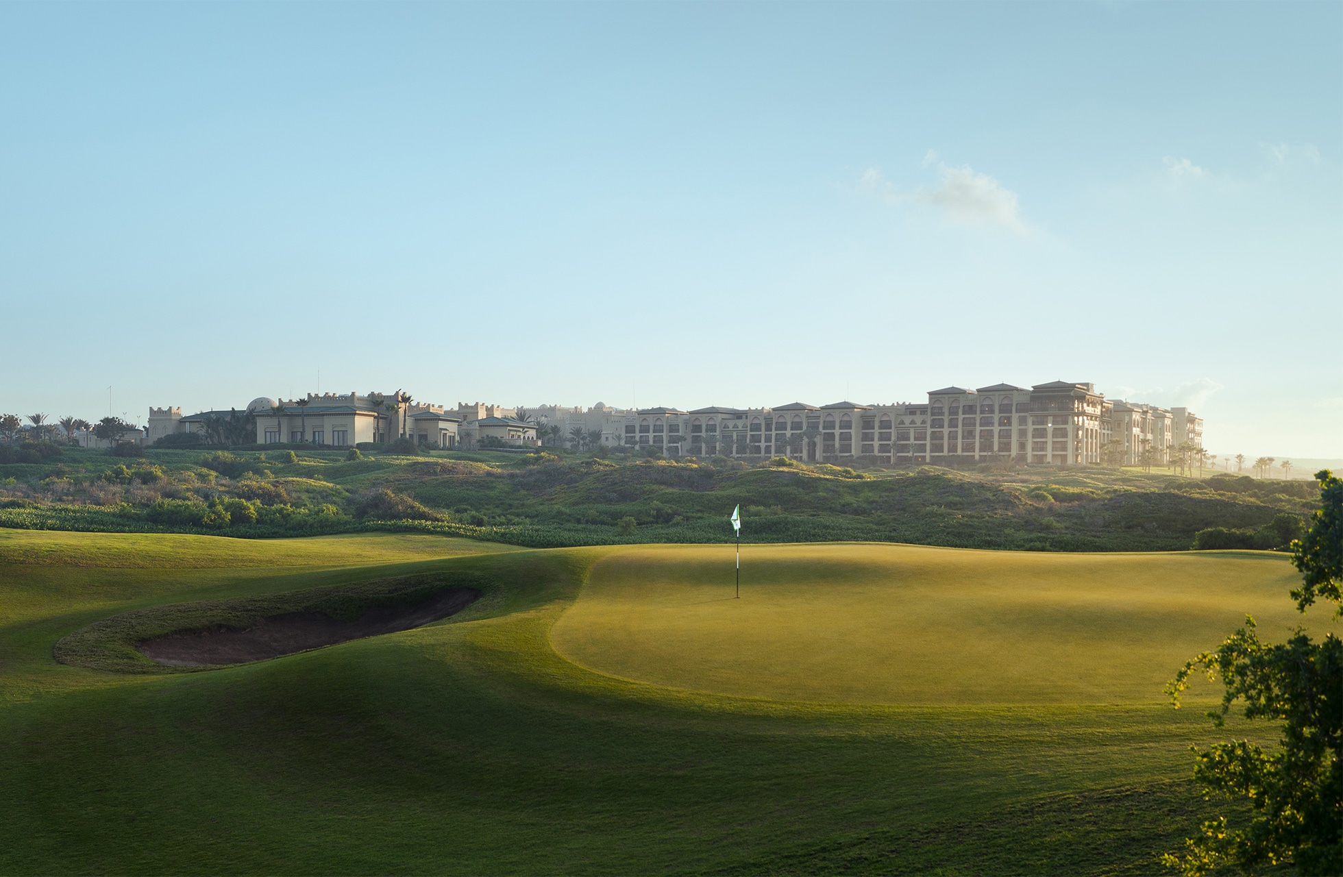 golfgreen med flagg og bunker i forgrunnen, mykt bølgende fairwayer og et stort hotellkompleks synlig i bakgrunnen i dagslys