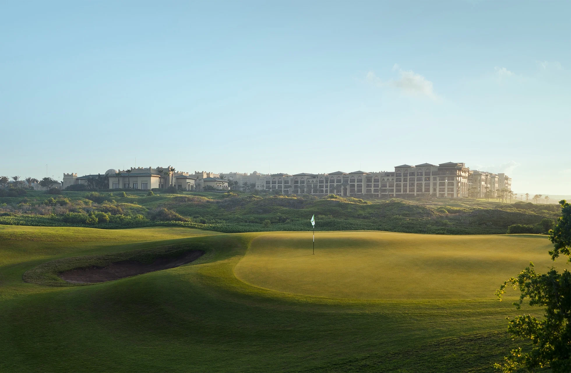 golfgreen med flagg og bunker i forgrunnen, mykt bølgende fairwayer og et stort hotellkompleks synlig i bakgrunnen i dagslys