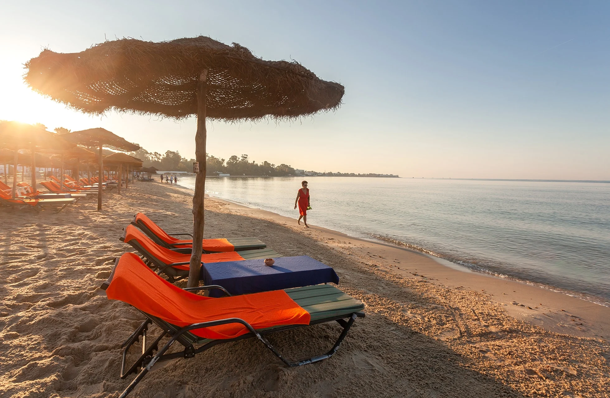 sandstrand med rekke av solsenger og stråparasoller, en person går langs vannkanten, rolig sjø og lav sol i varmt kveldslys